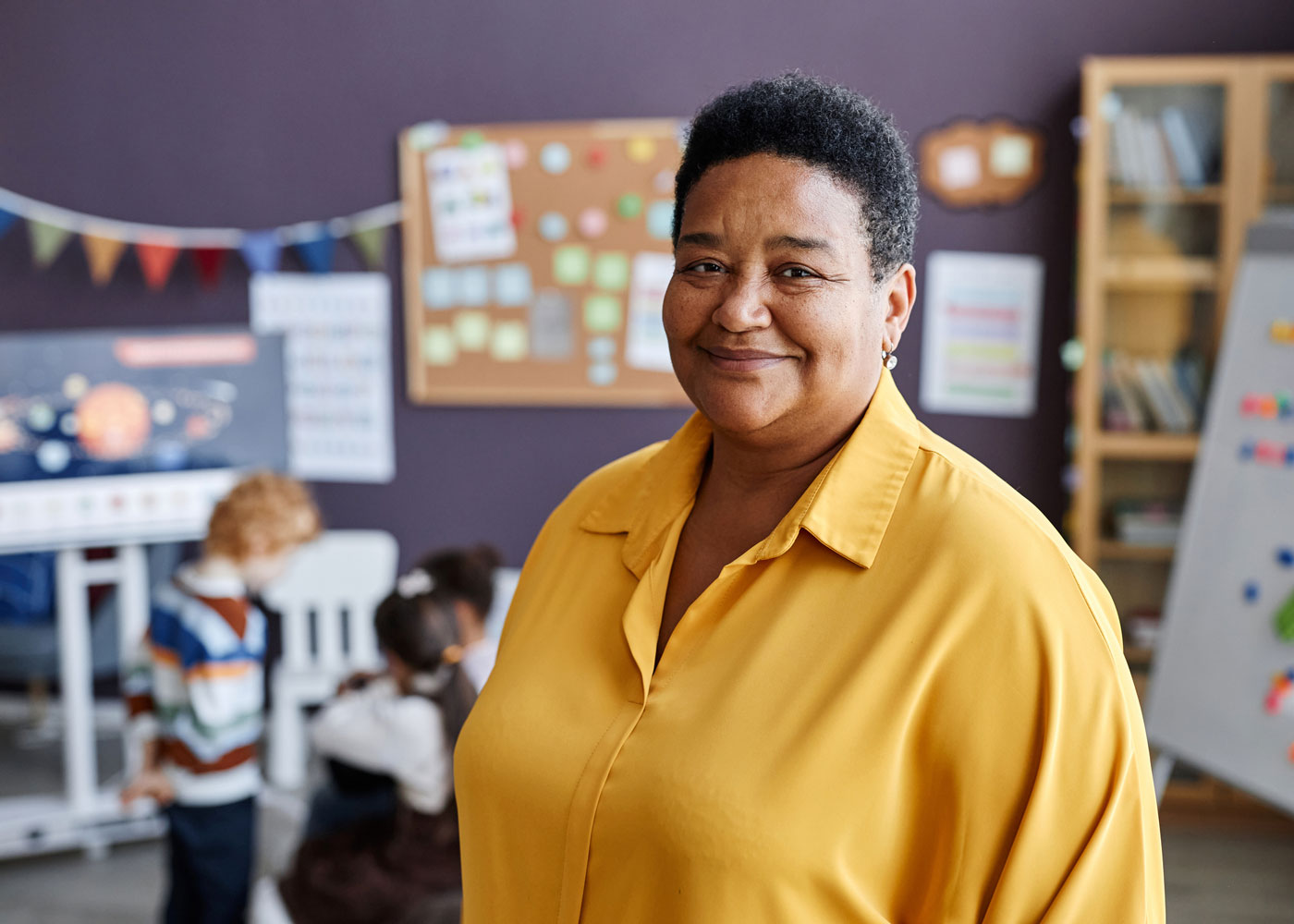 Female Teacher in a yellow shirt with kids in her classroom behind her.