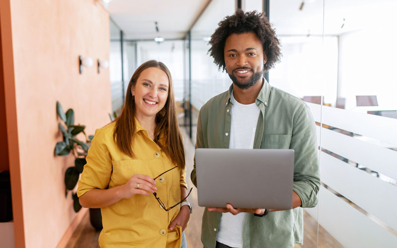 Two young coworkers, one female and one male, standing in their office smiling.