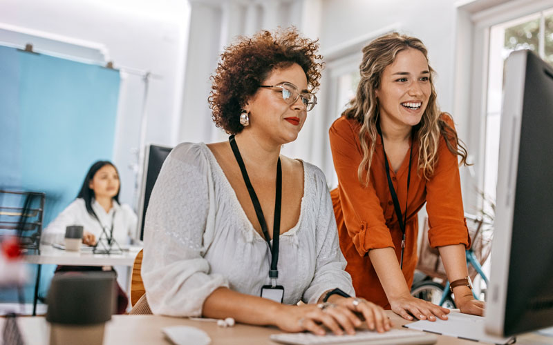 Two young female coworkers at a computer, one standing and one sitting.